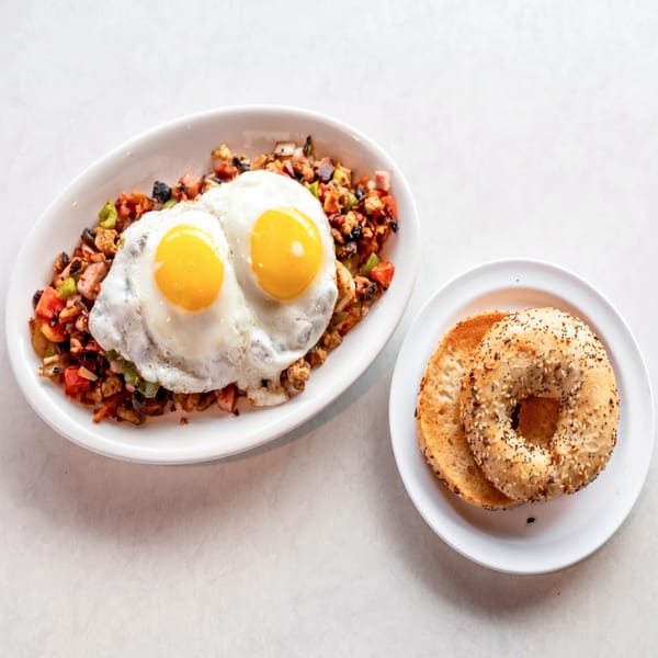 Breakfast hash with two fried eggs and a toasted everything bagel on a white table.
