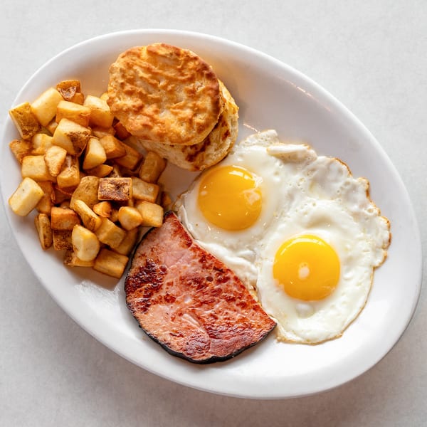Breakfast plate with two fried eggs, a slice of ham, home fries, and two biscuits.