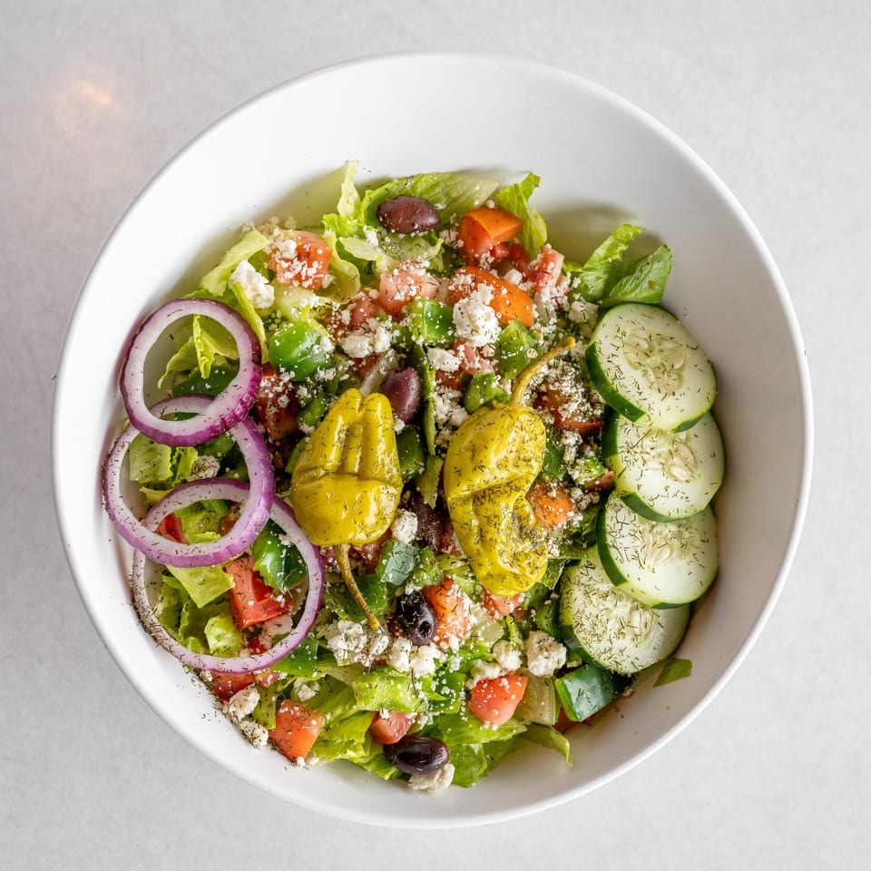 A vibrant Greek salad in a white bowl, featuring lettuce, tomatoes, cucumbers, red onions, feta, olives, and pepperoncini.