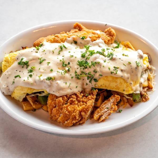 A white plate holds a chicken fried steak omelet covered in gravy and parsley.