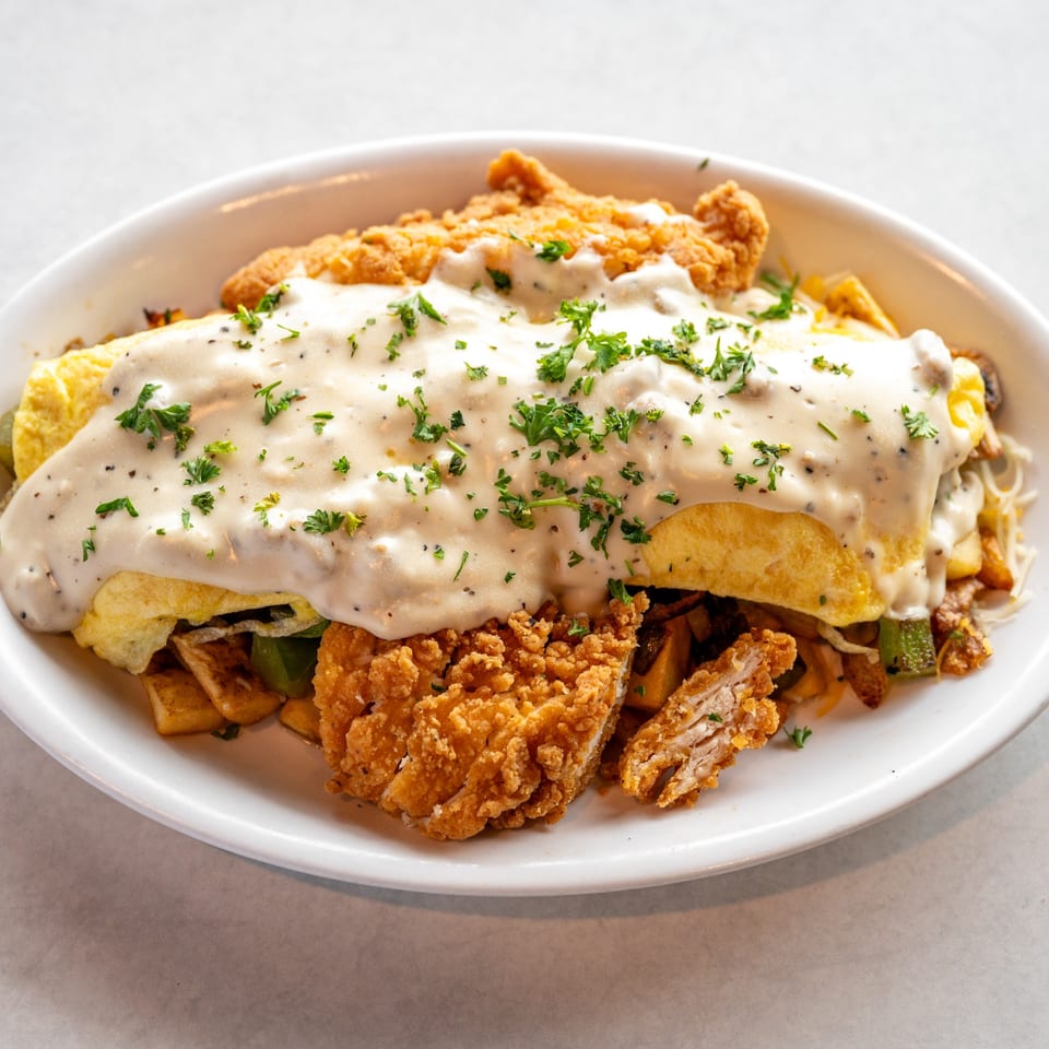 A white plate holds a chicken fried steak omelet covered in gravy and parsley.
