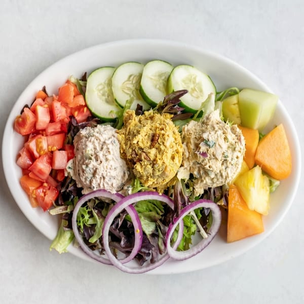 A salad with three scoops of chicken salad, tomatoes, cucumbers, red onions, and fruit.