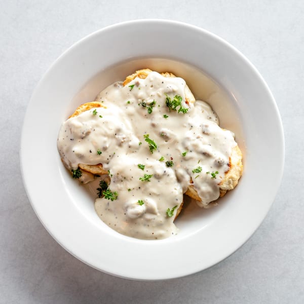 Biscuits and gravy in a white bowl, topped with fresh parsley, on a light background.